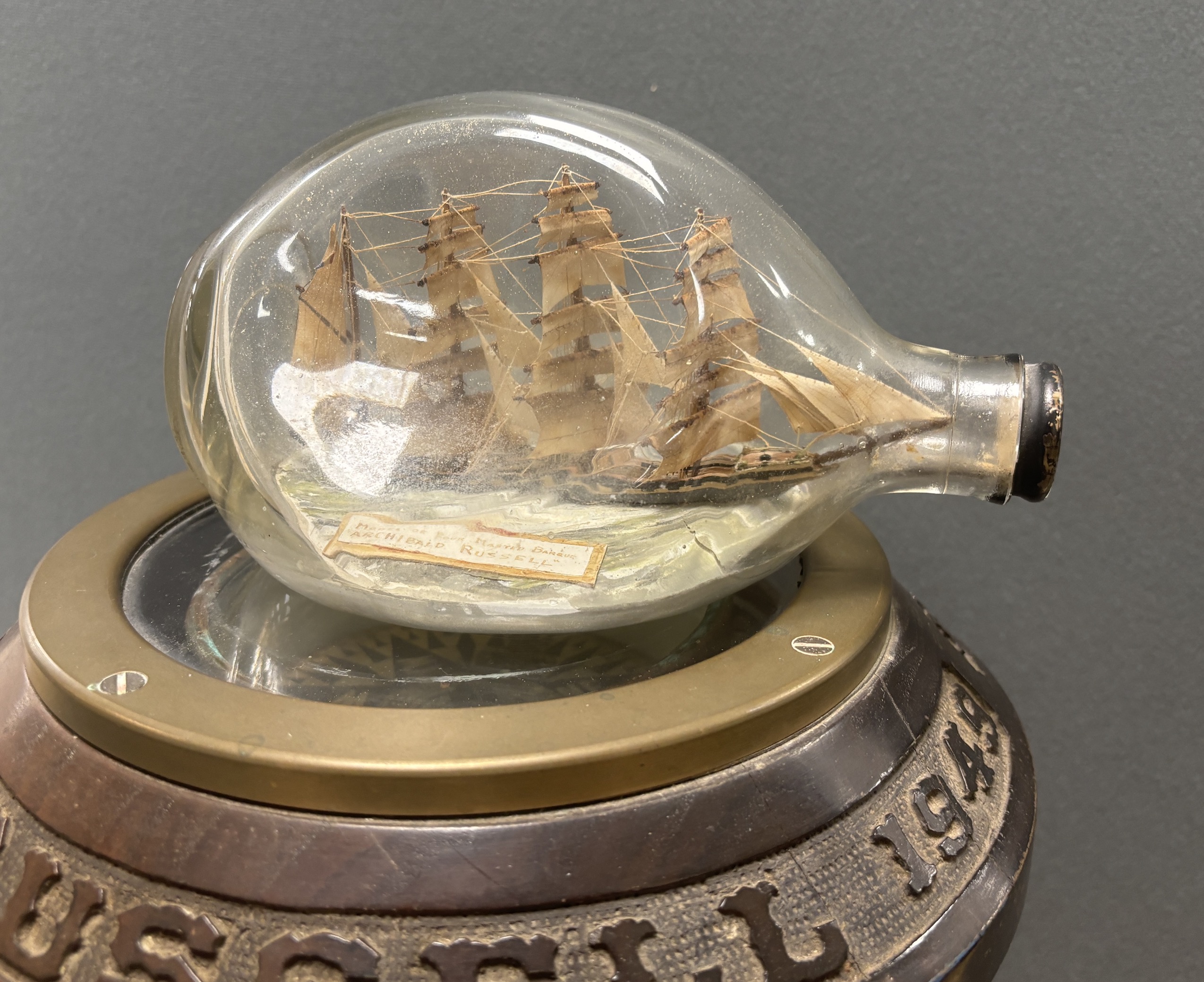 A ship’s compass in a gimbal, believed to be from the tall ship The Archibald Russell, mounted on a floor standing mahogany stand with carved attribution around the edge, together with a ship in a bottle of the barque in